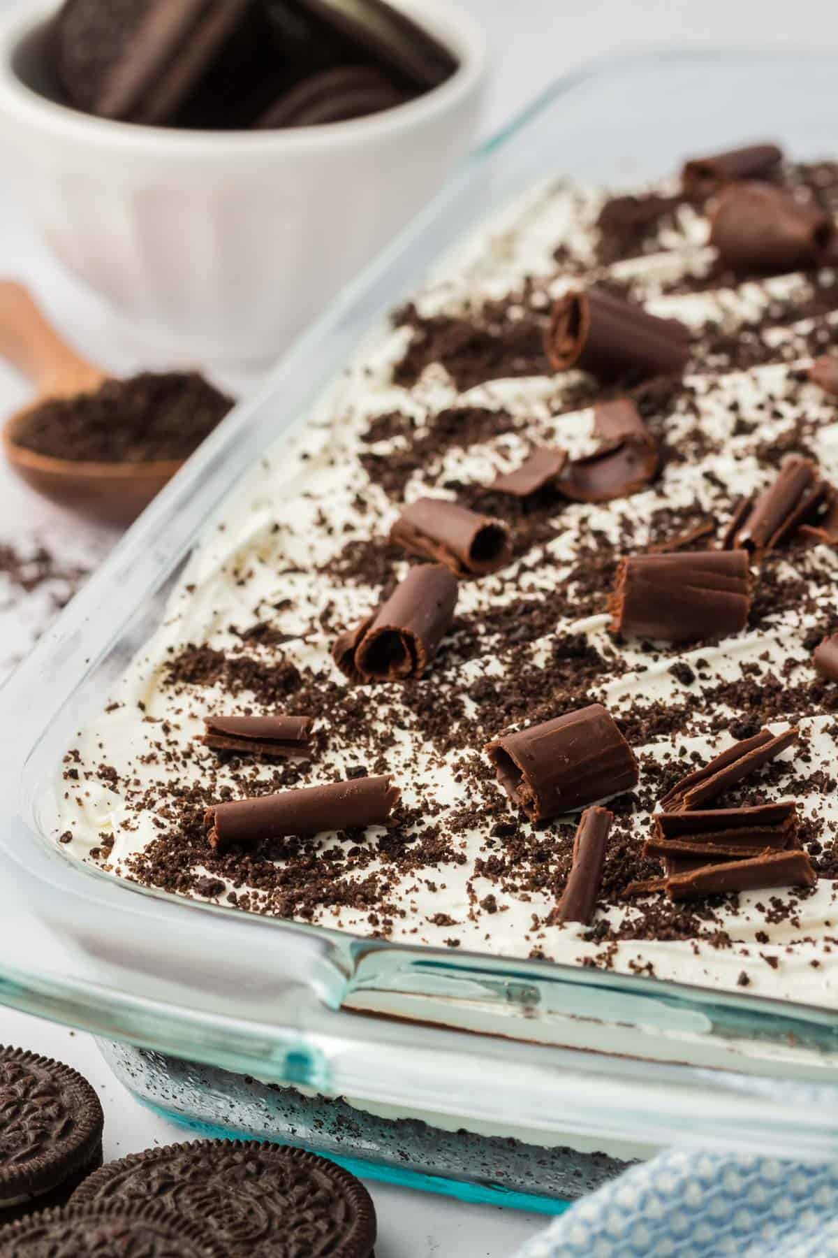 a close up of the top of a pan of chocolate lasagna topping with chocolate shavings and oreo crumbs then surrounded by whole oreos and a spoon full of oreo crumbs