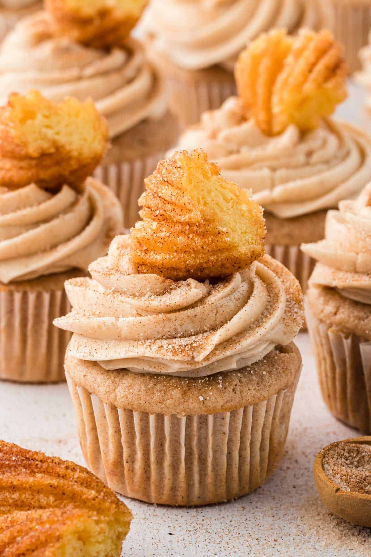 churro cupcakes arranged around each other with churro pieces on top, a small wooden spoon of cinnamon sugar and on churro in the corner