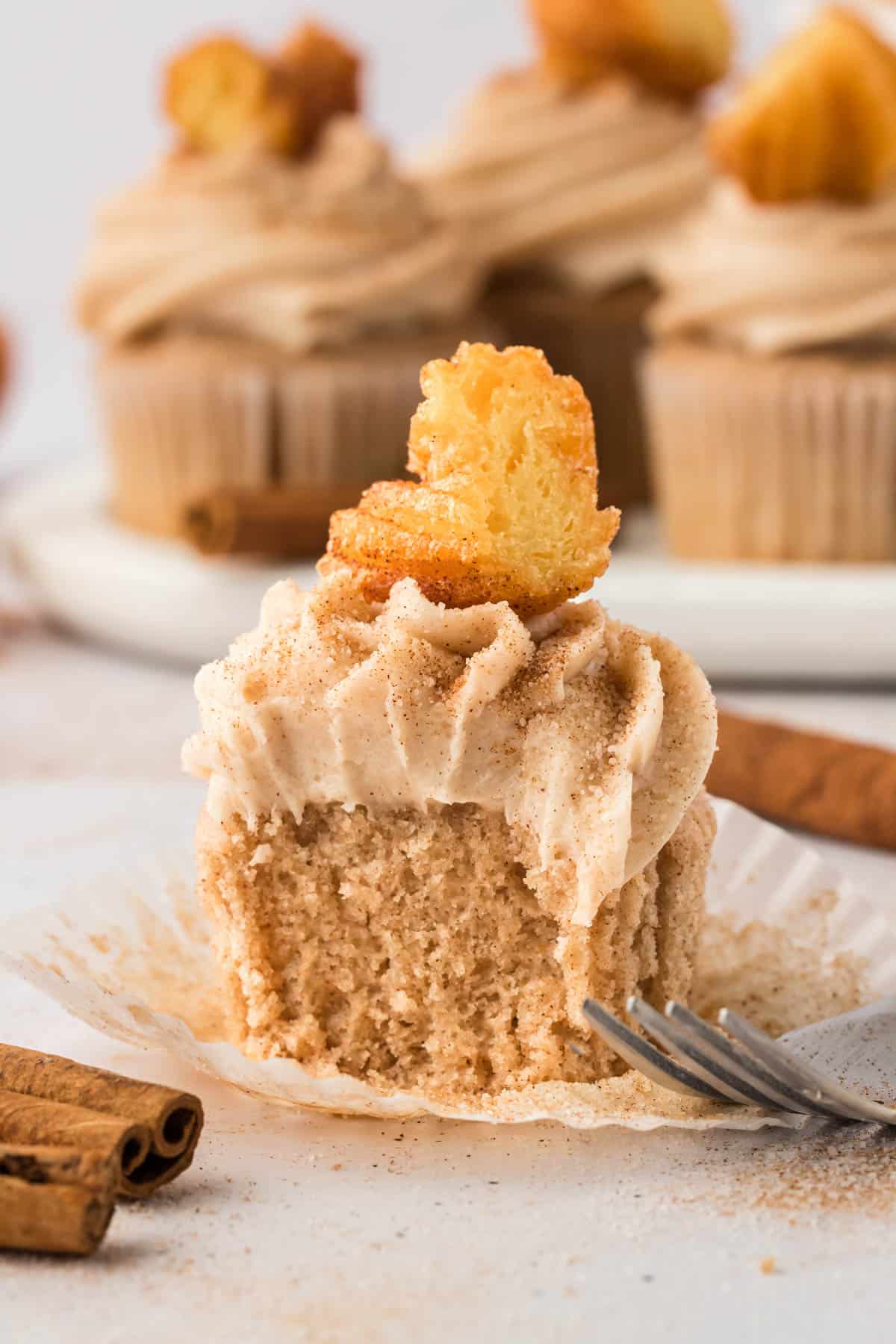 a churro cupcake in an open muffin liner with a fork laying by it and a bit taken out of the cupcake by a fork, surrounded by cinnamon sticks and a dust of cinnamon sugar and more cupcakes in the background
