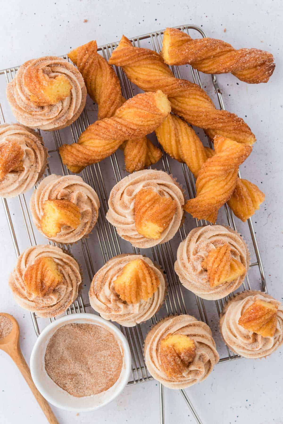 churro cupcakes topped with churro pieces on top of a wire rack surrounded by churros, a wooden spoon and a white bowl of cinnamon sugar