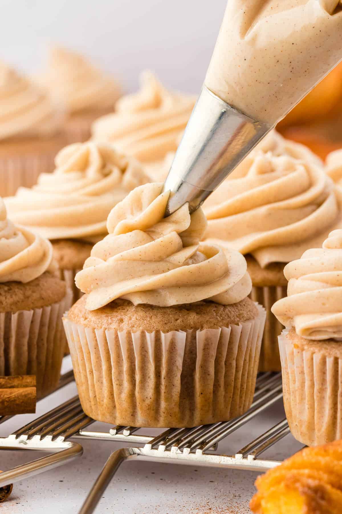 cinnamon cream cheese frosting being piped on top of a cupcake that is sitting on a wire cooling rack with more cupcakes that have already been iced