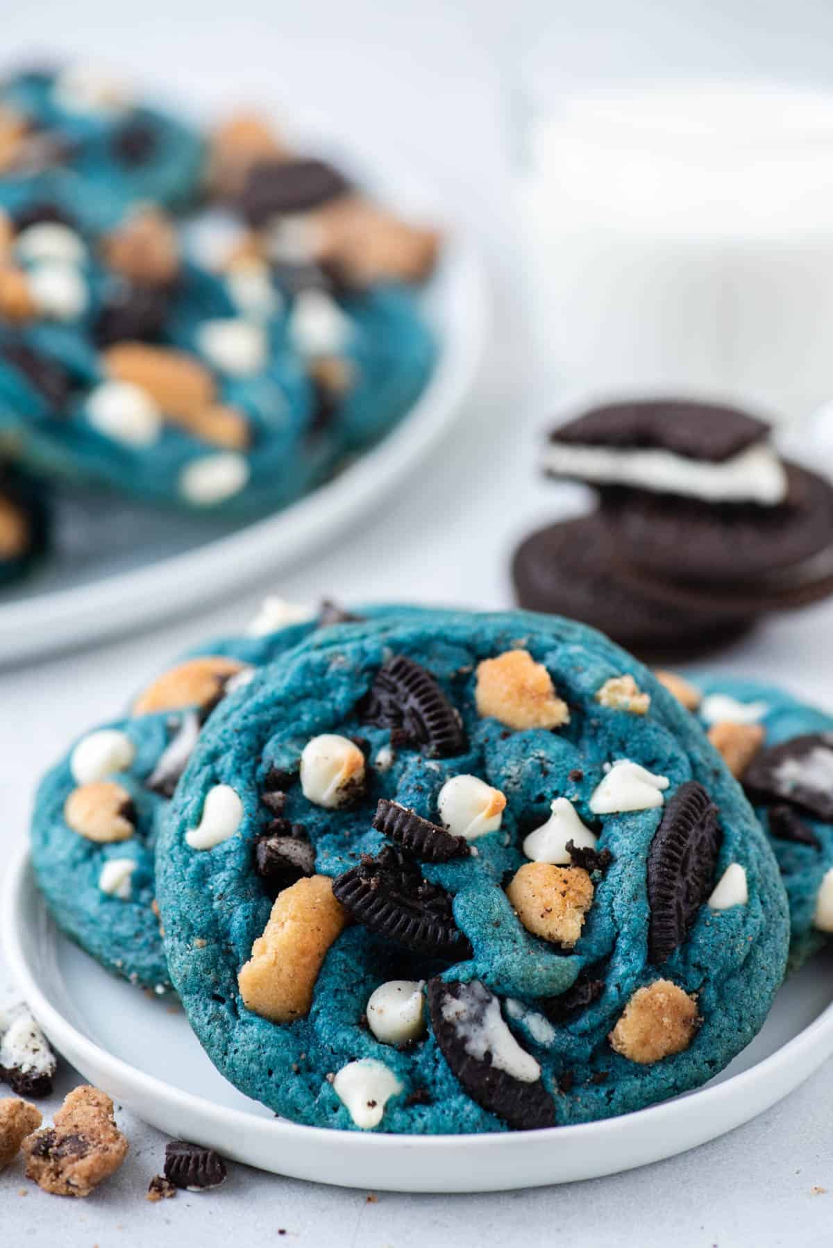 a white plate with three cookie monster cookies on it surrounded by cookie pieces with oreos and more cookies in the background