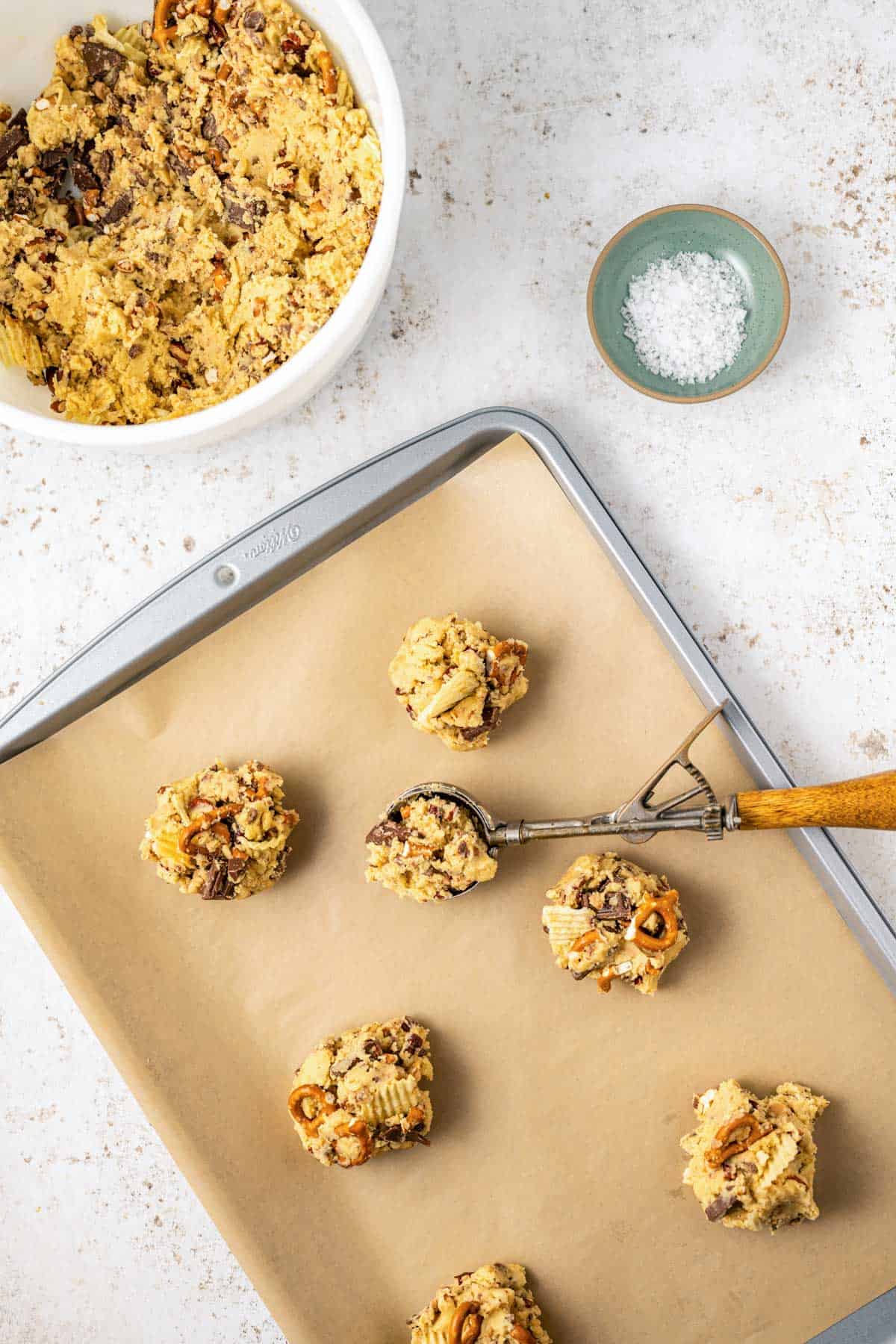 a baking sheet lined with parchment paper with kitchen sink cookie dough balls lined on the sheet, with one still in the cookie scoop, a small bowl of salt on the side and a larger bowl of cookie dough beside that