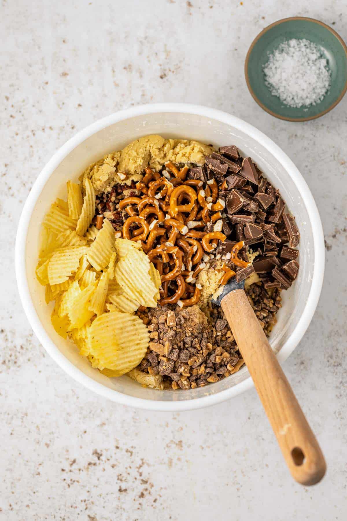 a white bowl full of kitchen sink cookie ingredients with piles of potato chips, toffee bits, pretzels and chocolate chunks on top, with a wooden spatula stuck inside the bowl and a small bowl of salt on the side
