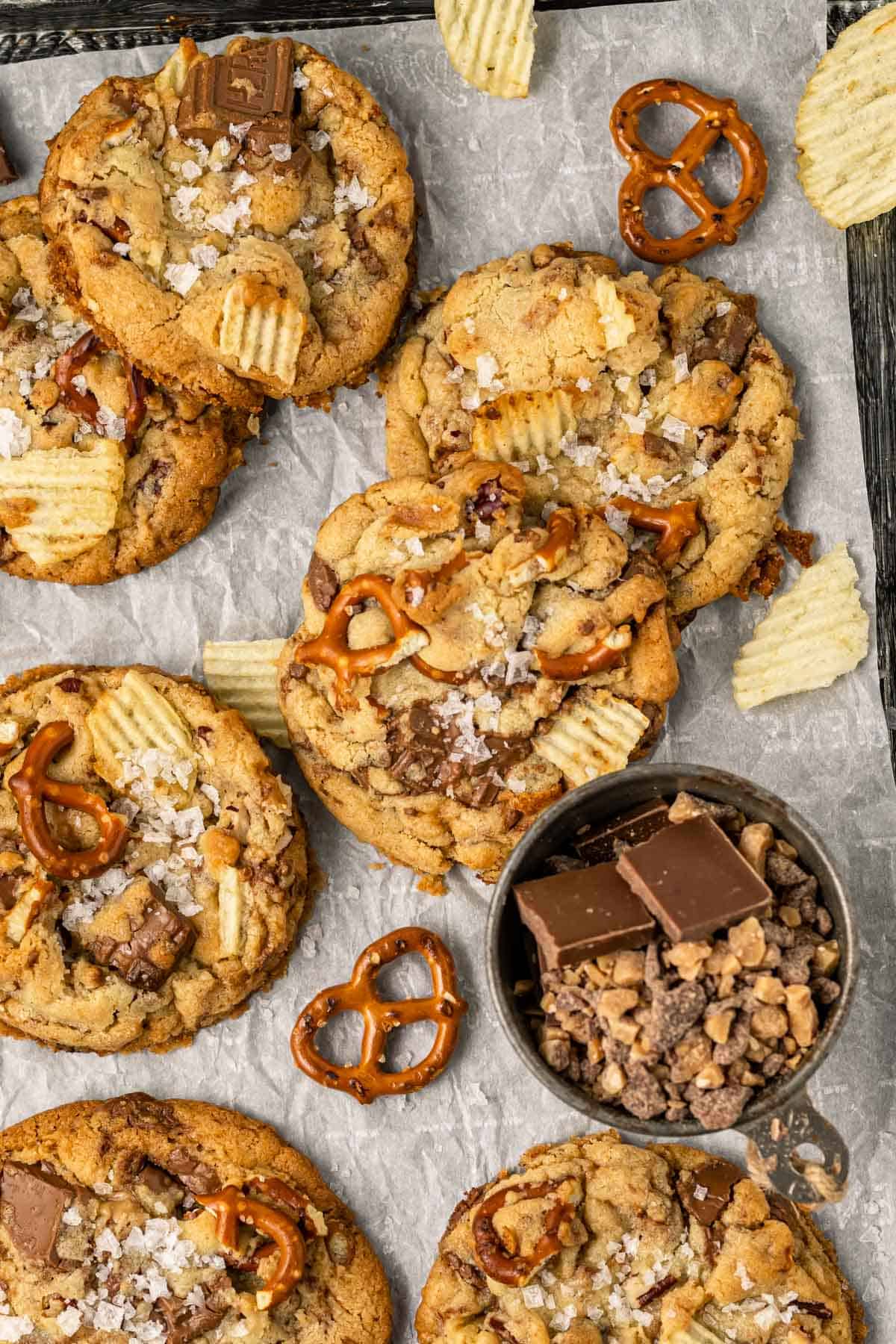 a sheet pan lined with parchment paper with kitchen sink cookies arranged on top surrounded by potato chips, pretzels, chunks of chocolate, and a small black dish of toffee bits
