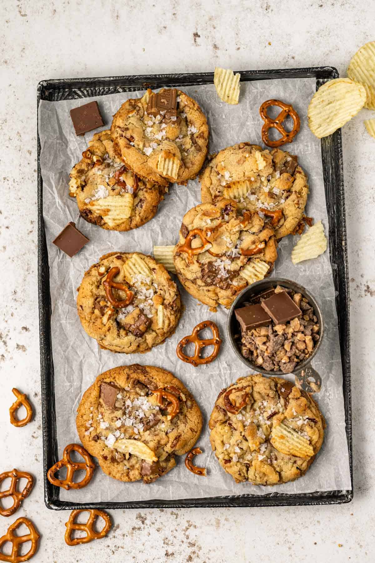 a sheet pan lined with parchment paper with kitchen sink cookies arranged on top surrounded by potato chips, pretzels, chunks of chocolate, and a small black dish of toffee bits