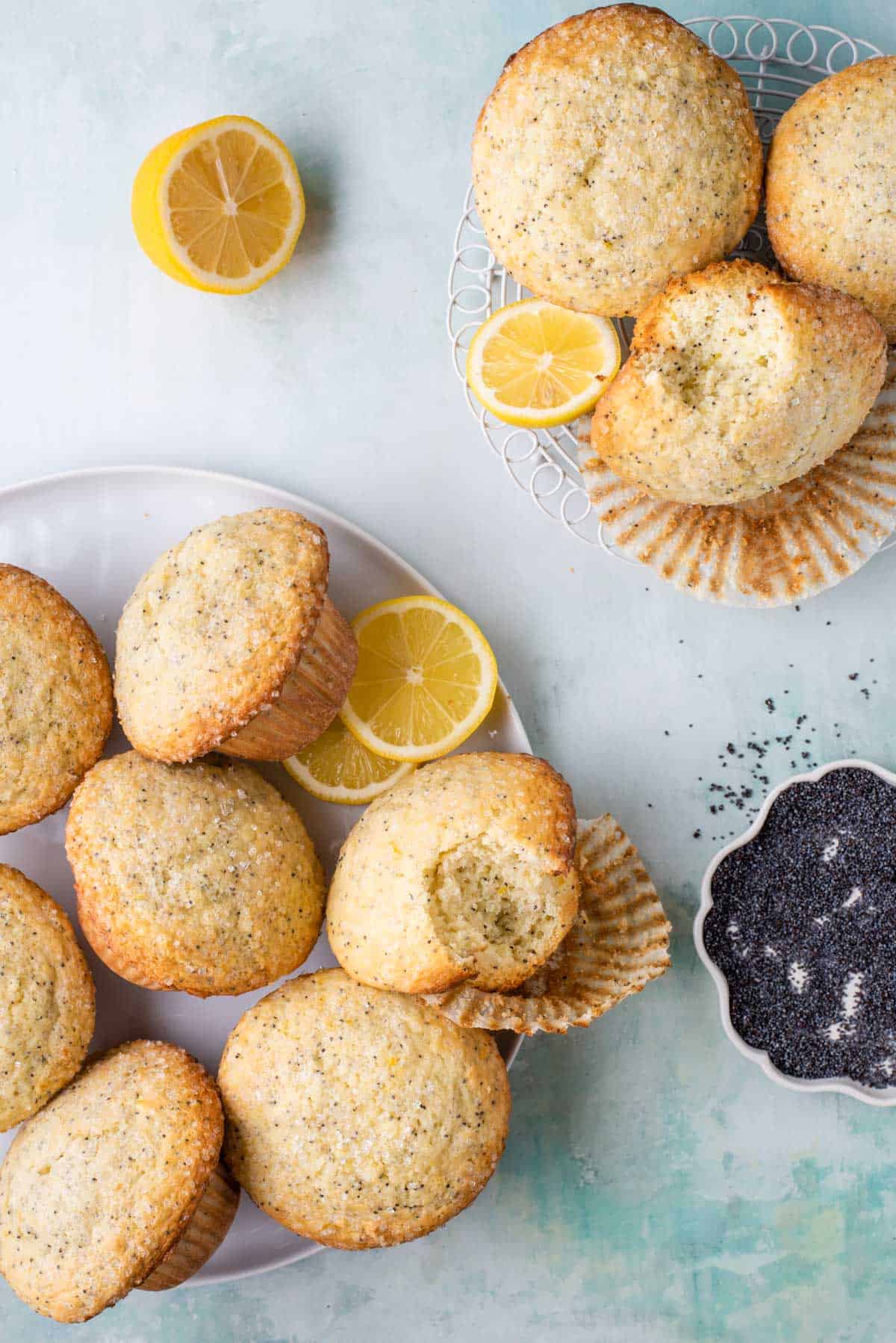 two plates of lemon poppy seed muffins piled up, some partially eaten, arranged with lemon slices and a bowl of poppy seeds