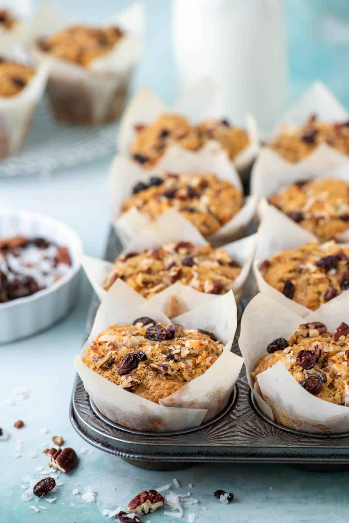 morning glory muffins in muffin liners inside a muffin pan surrounded by nuts, raisins and coconut shreds and more muffins in the background