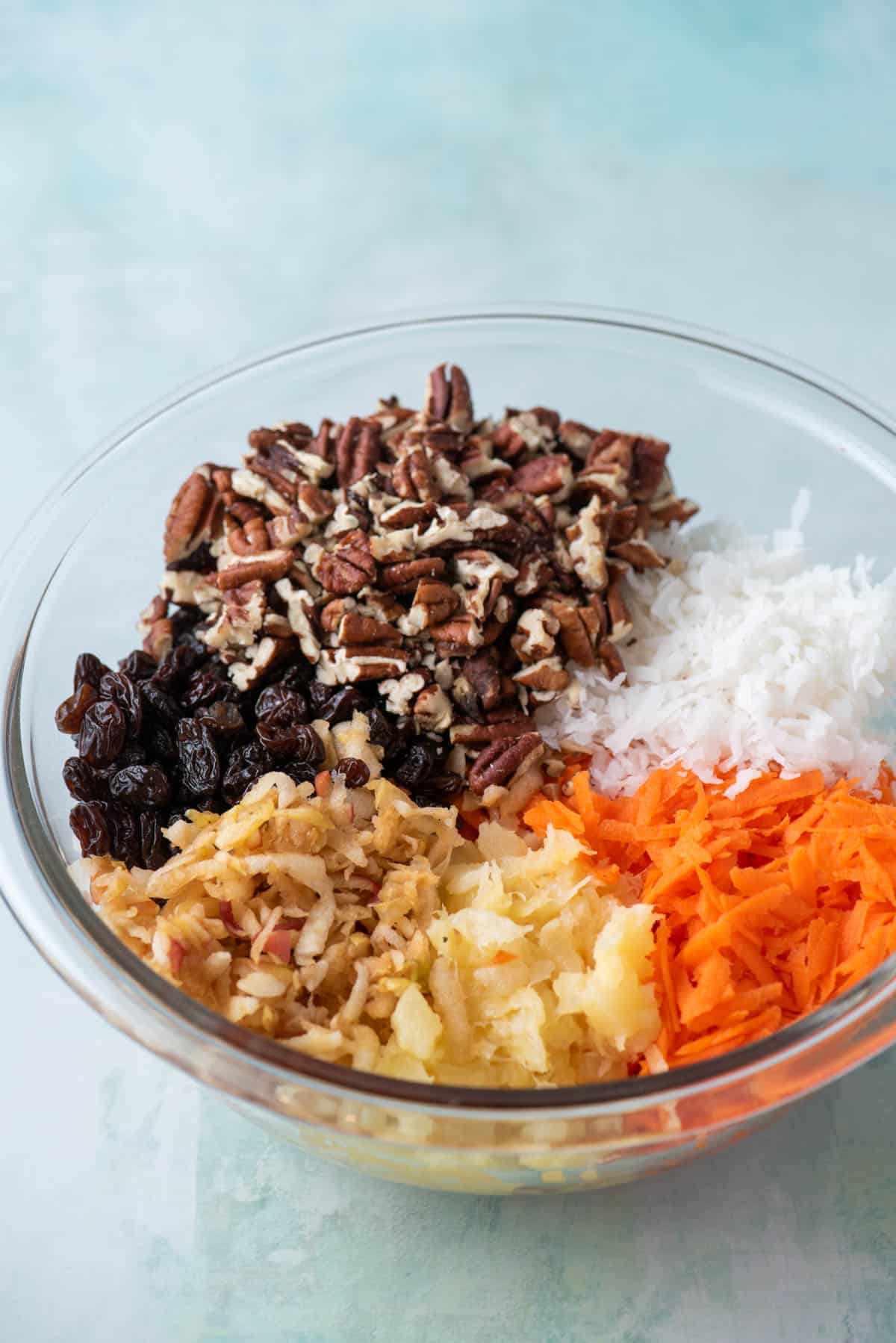 ingredients for morning glory muffins in a glass bowl including raisins, walnuts, shredded apple, crushed pineapple, shredded carrots and shredded coconut