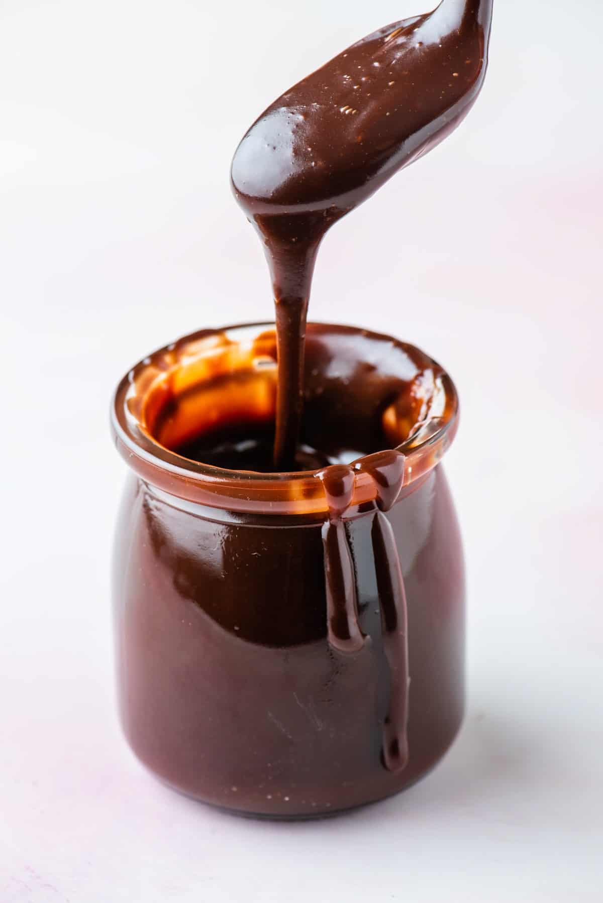 a jar of hot fudge sauce with a spoon above it full of sauce that is dripping down and sauce dripping down the side of the jar