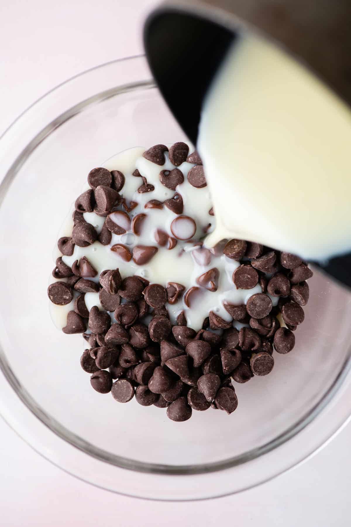 a mixture of heavy cream and sugar being poured over a glass bowl of chocolate chips to make homemade hot fudge sauce
