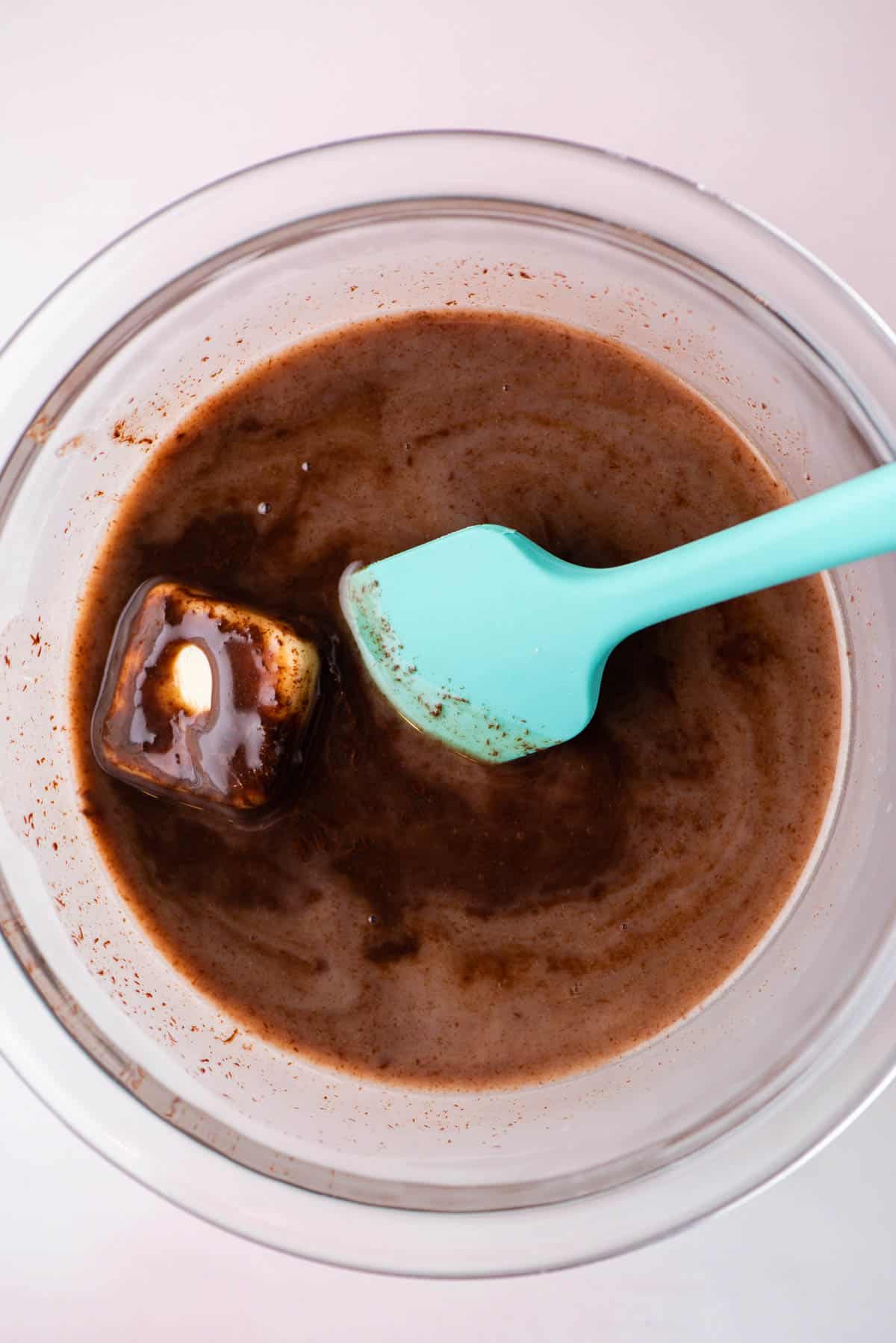 hot fudge sauce being made in a glass bowl by mixing in butter to the rest of the ingredients using a teal spatula