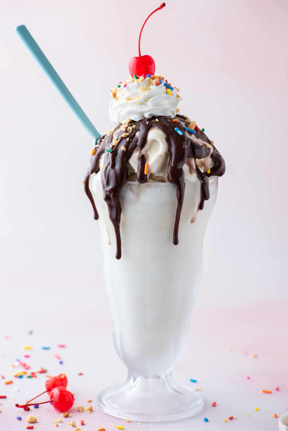 a hot fudge sunday in a tall glass surrounded by sprinkles, bits of peanuts and a couple maraschino cherries with a blue spoon stuck into the side of the sundae