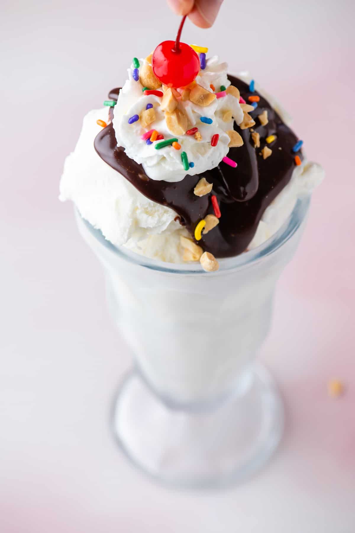 a hot fudge sundae in a tall glass topped with sprinkles, peanuts and cool whip with a cherry being placed on top