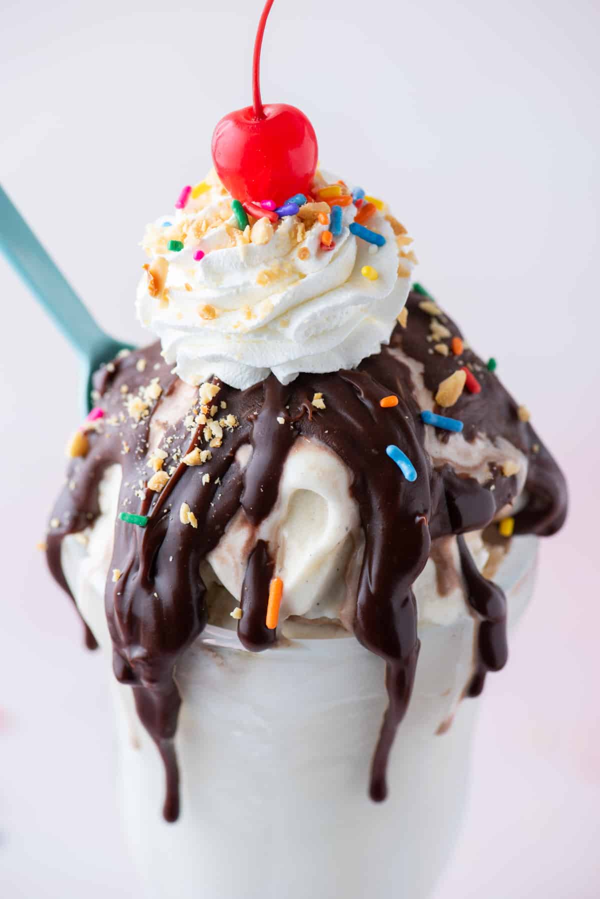 close up of a hot fudge sundae topped with sprinkles, peanuts, cool whip and a cherry on top with a blue spoon stuck into the side