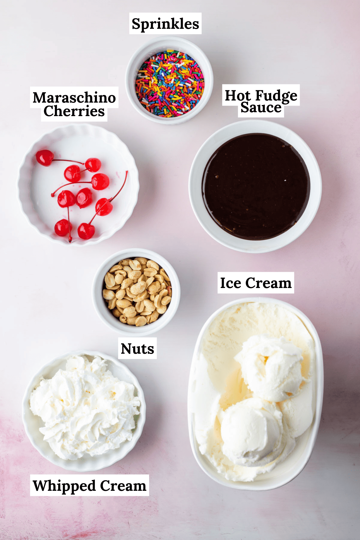 the ingredients for a hot fudge sundae laid out including a bowl of sprinkles, a bowl of hot fudge sauce, a bowl of maraschino cherries, a bowl of peanuts, a bowl of whipped cream and a bowl of vanilla ice cream
