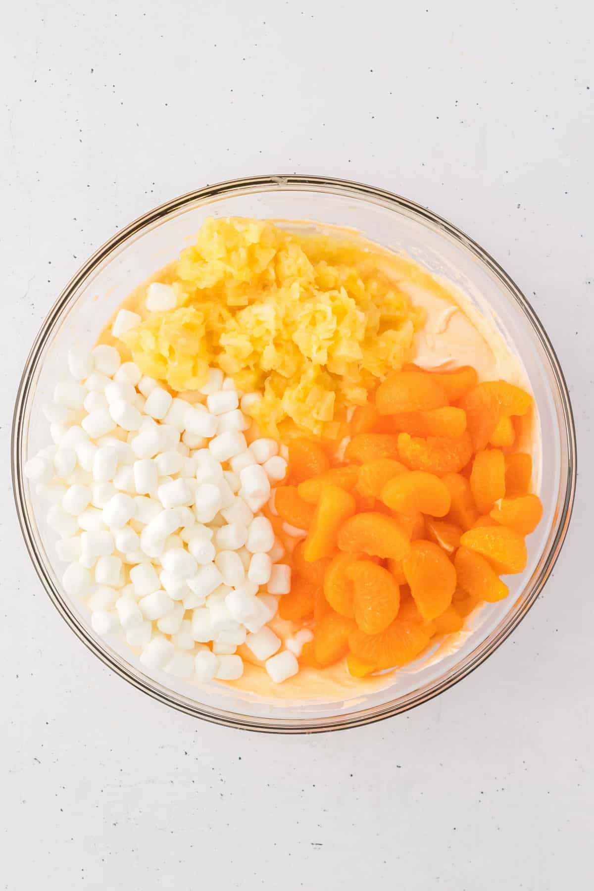 ingredients for orange fluff salad in a bowl with mini marshmallows, crushed pineapple and mandarin oranges on top that haven't been stirred in yet