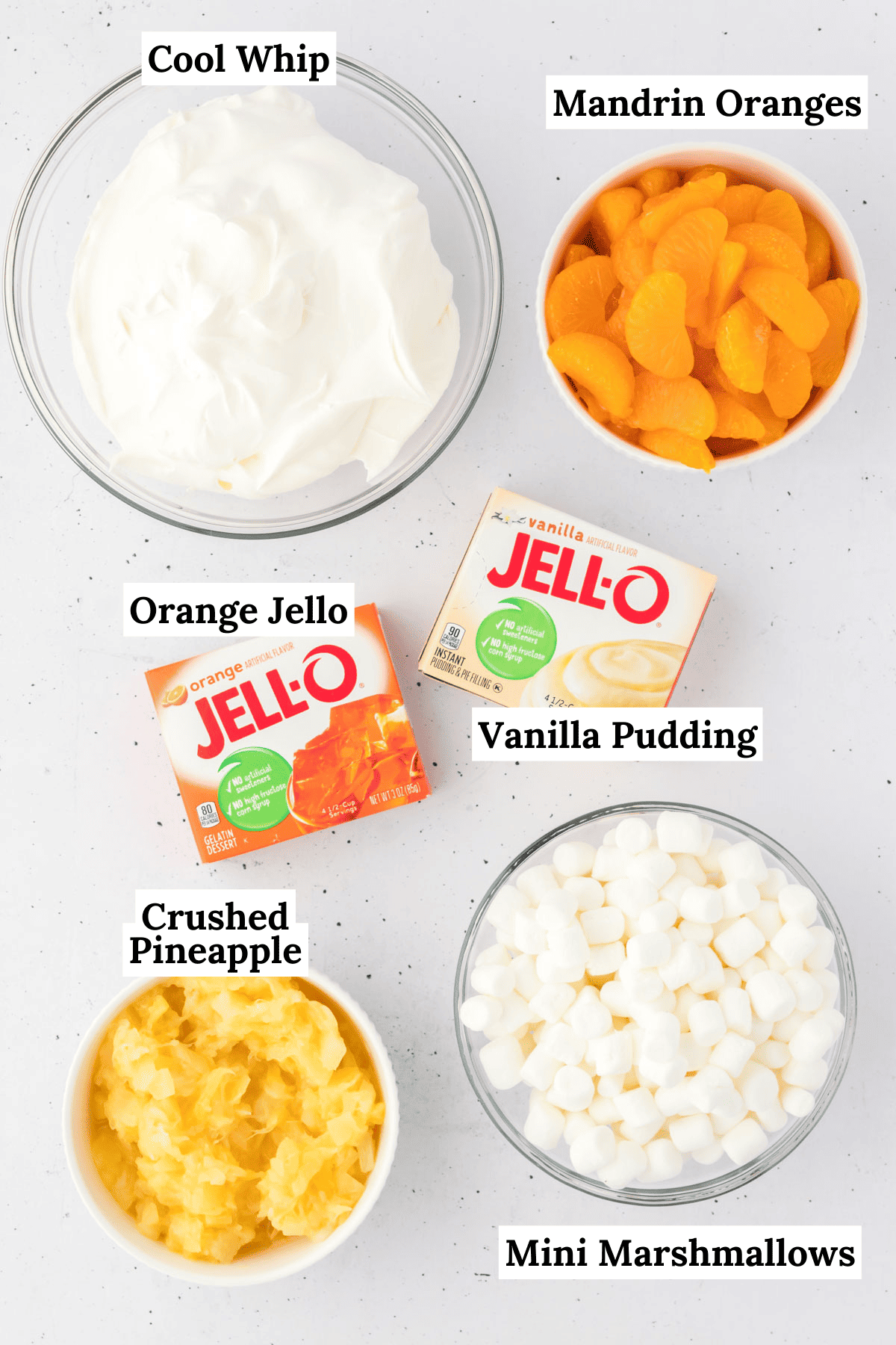 the ingredients for orange fluff salad layed out on a counter including orange jello mix, vanilla pudding mix, a bowl of mini marshmallows, a bowl of crushed pineapple, and a bowl of cool whip