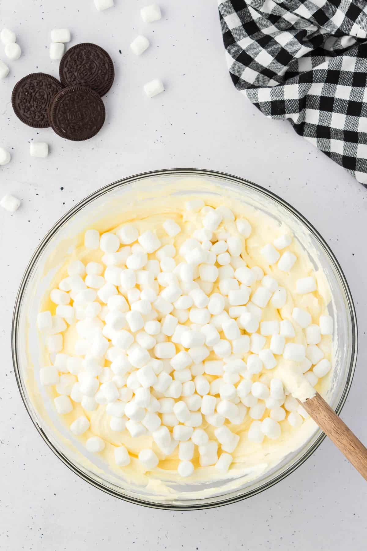 vanilla pudding mixture topped with mini marshmallows in a glass bowl, surrounded by oreos, mini marshmallows and a black and white checkered kitchen towel