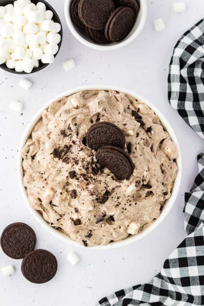 oreo fluff topped with oreo crumbs and two whole oreos in a glass bowl surrounded by oreos, mini marshmallows and a black and white checkered kitchen towel