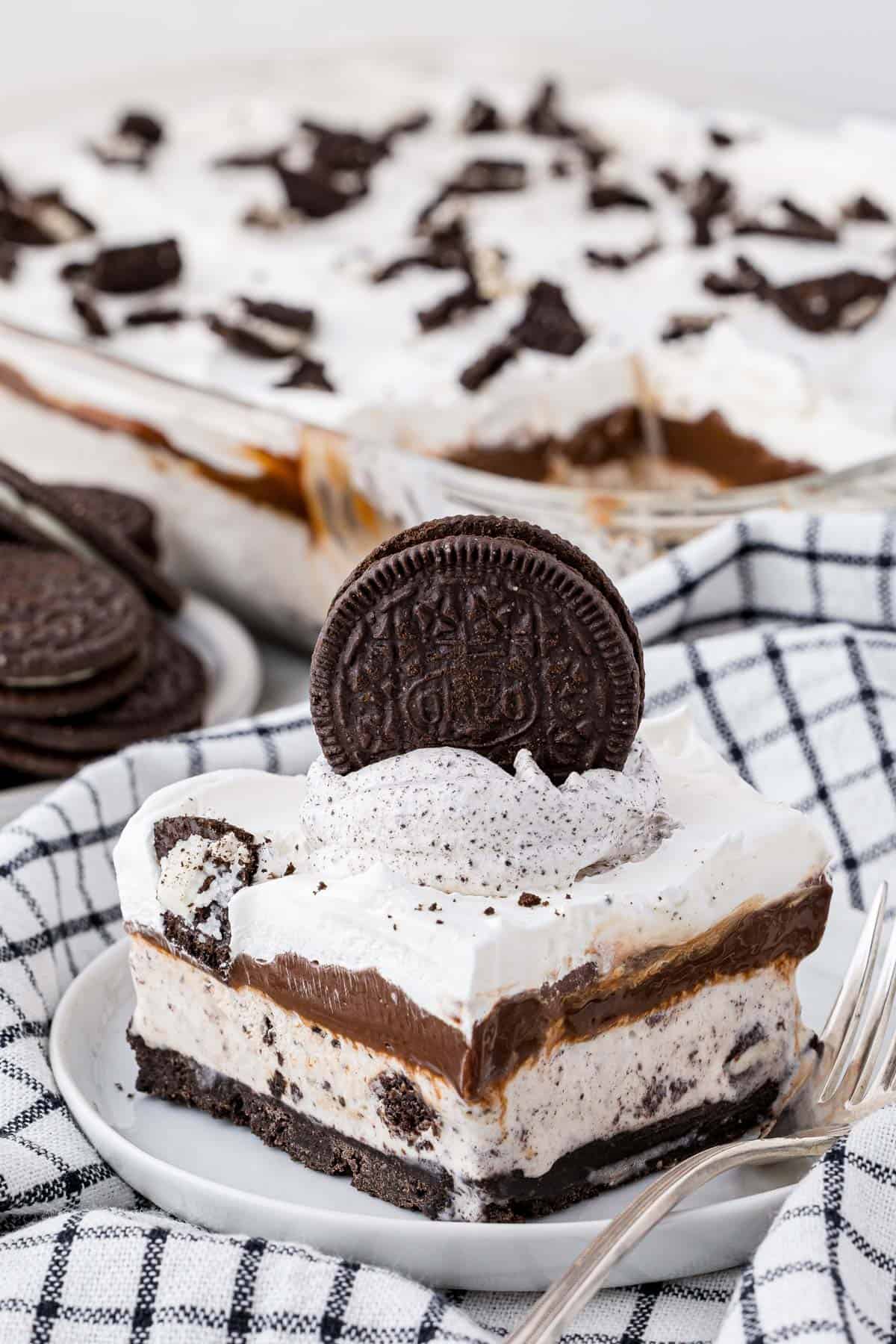 a slice of oreo ice cream cake on a white plate with a fork, sitting on top of a black and white checkered kitchen towel with more ice cream cake and oreos in the background