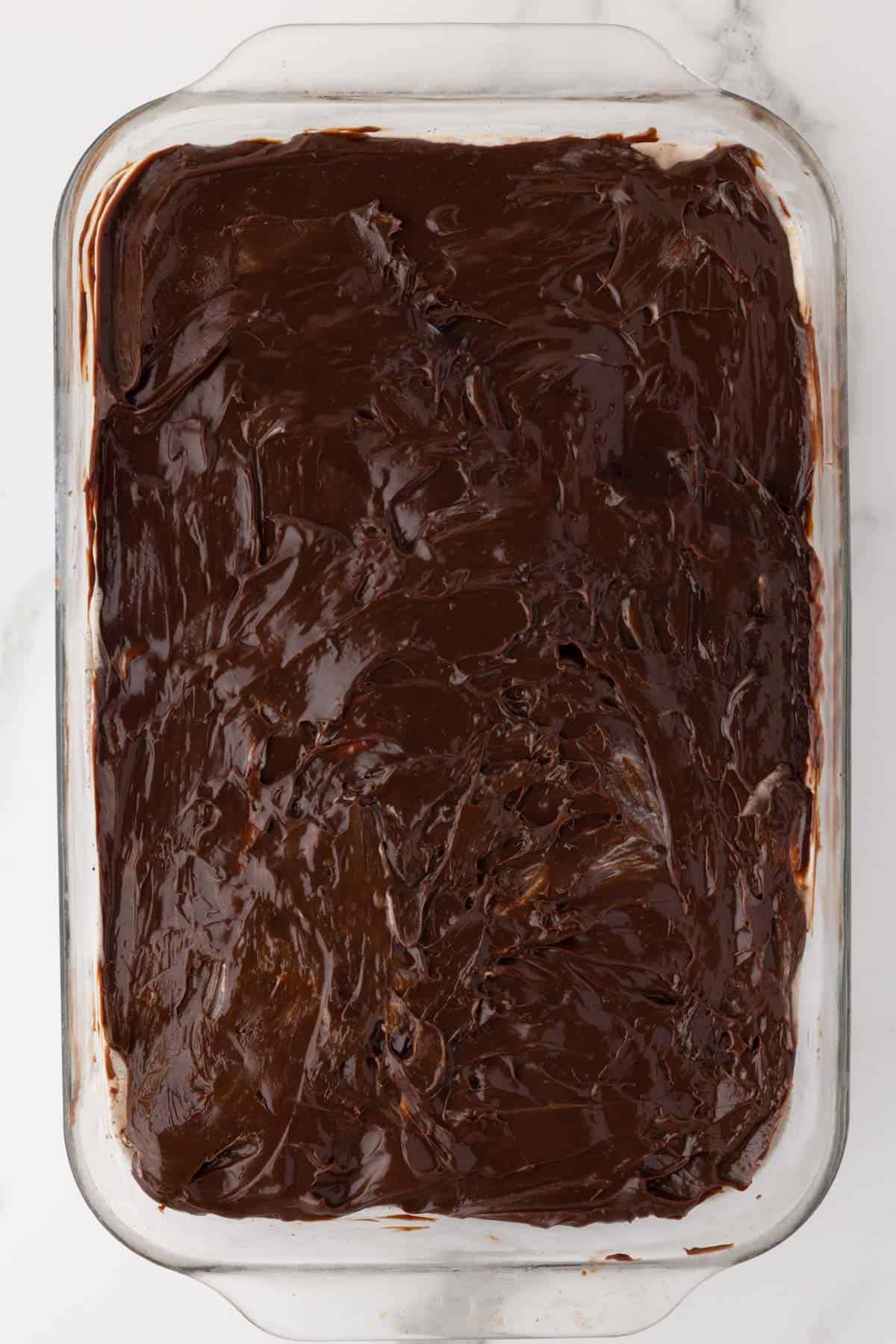 layers of oreo ice cream cake being added to a baking dish with the layer of hot fudge showing on top