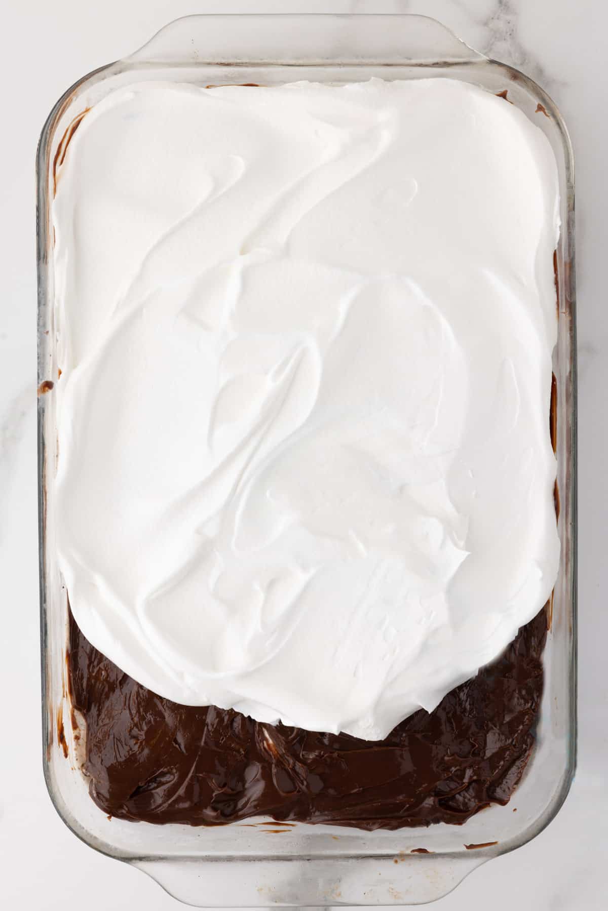 layers of oreo ice cream cake being added to a baking dish with the hot fudge layer showing and the cool whip layer half spread on top