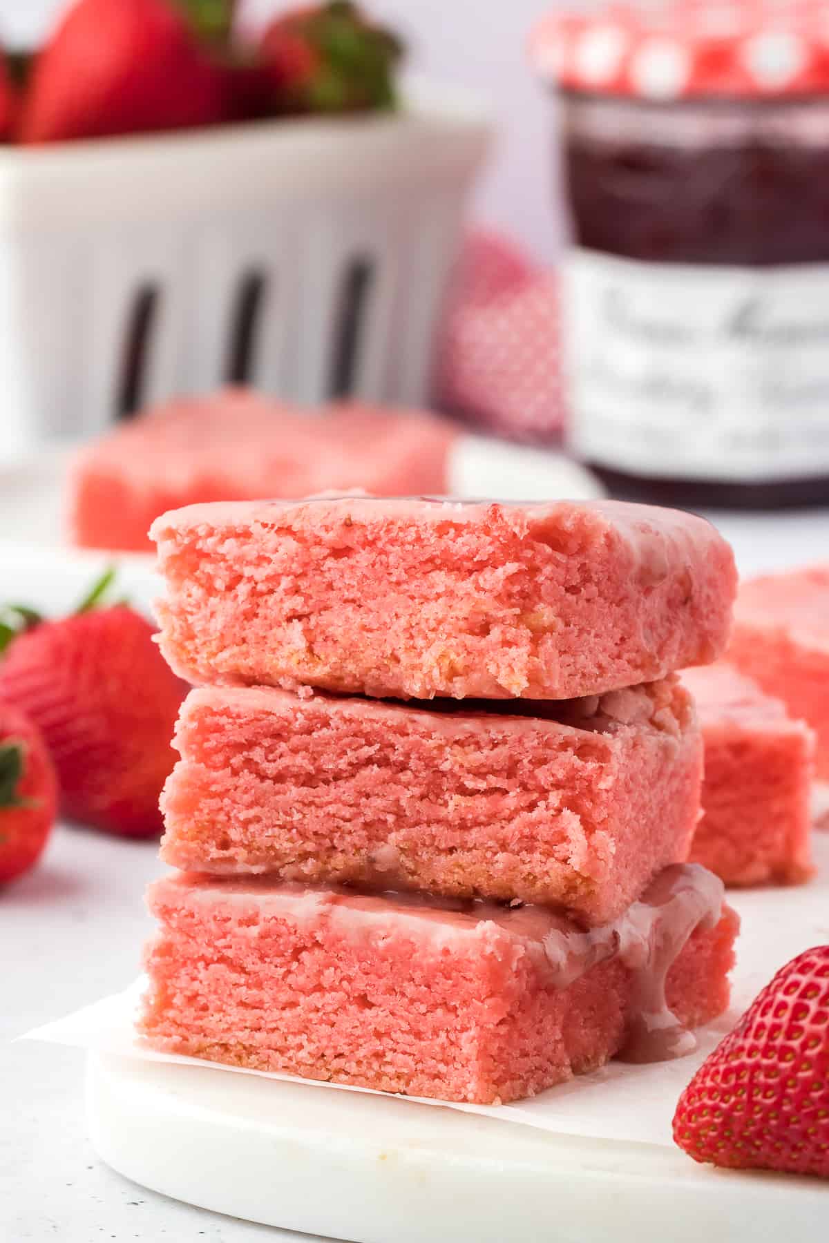 three strawberry brownies stacked surrounded by a red and white kitchen towel, fresh strawberries and strawberry preserves