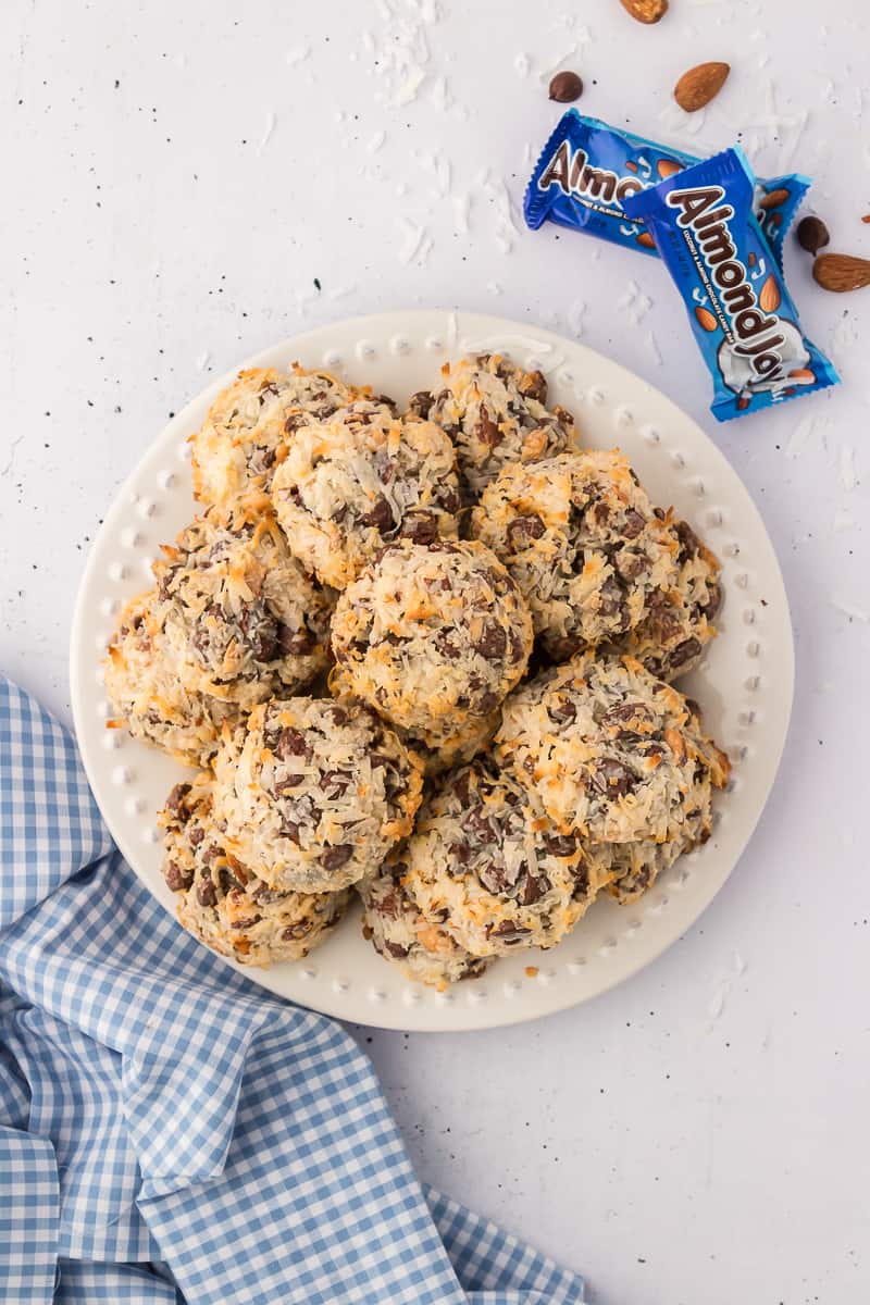 A pile of almond joy cookies on a white plate with two almond joy candy bars beside it and coconut flakes, almonds and chocolate chips sprinkled around
