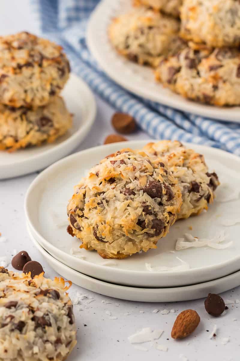 almond joy cookies on a white plate with coconut flakes and chocolate chips sprinkled around, a blue and white checkered kitchen towel and more plates with almond joy cookies on them in the background