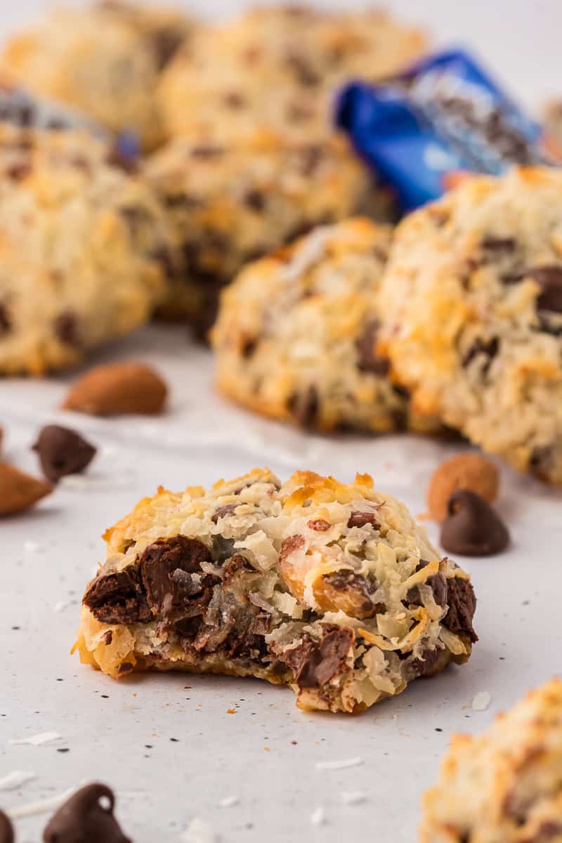 almond joy cookies arranged on a flat surface with chocolate chips and coconut flakes sprinkled around and a close up of one cookie with a bite of of it in the middle