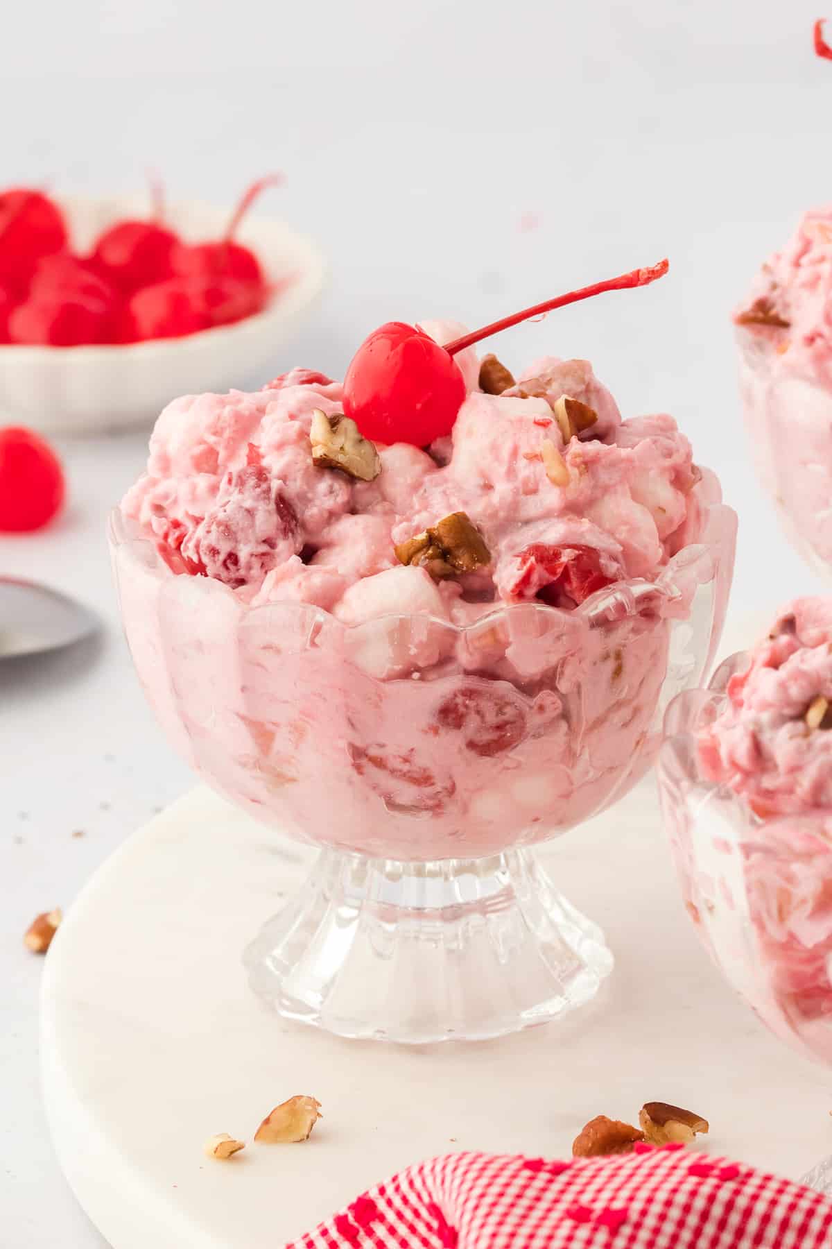 a glass bowl of cherry fluff topped with a maraschino cherry and surrounded by a bowl of cherries, more bowls of cherry fluff, and sprinkled nuts and a red and white towel around them