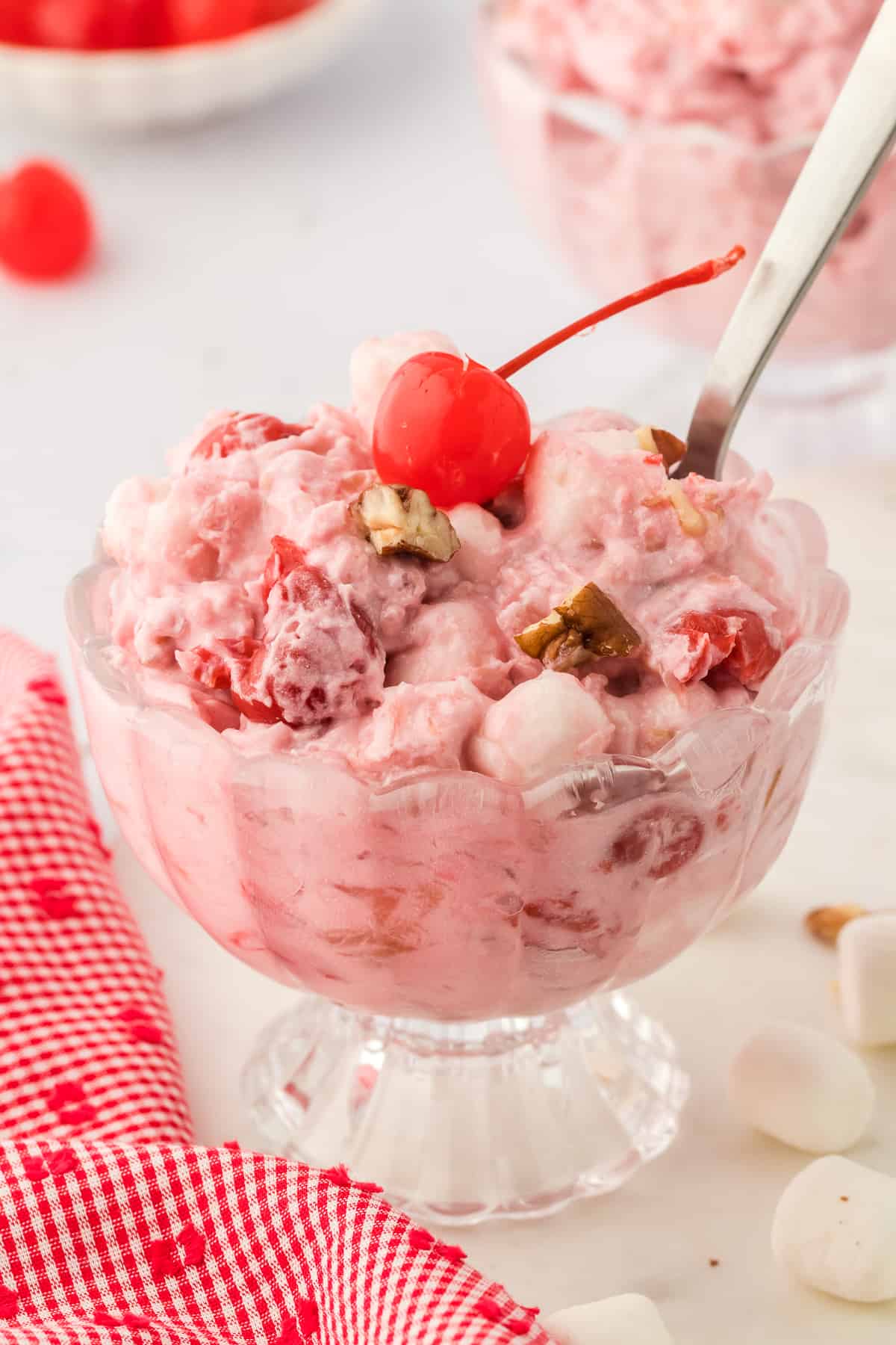 a glass bowl of cherry fluff topped with a maraschino cherry and a spoon stuck into it, with more fluff in the background and a bowl of cherries in the background, a red and white kitchen towel to the left and mini marshmallows sprinkled around