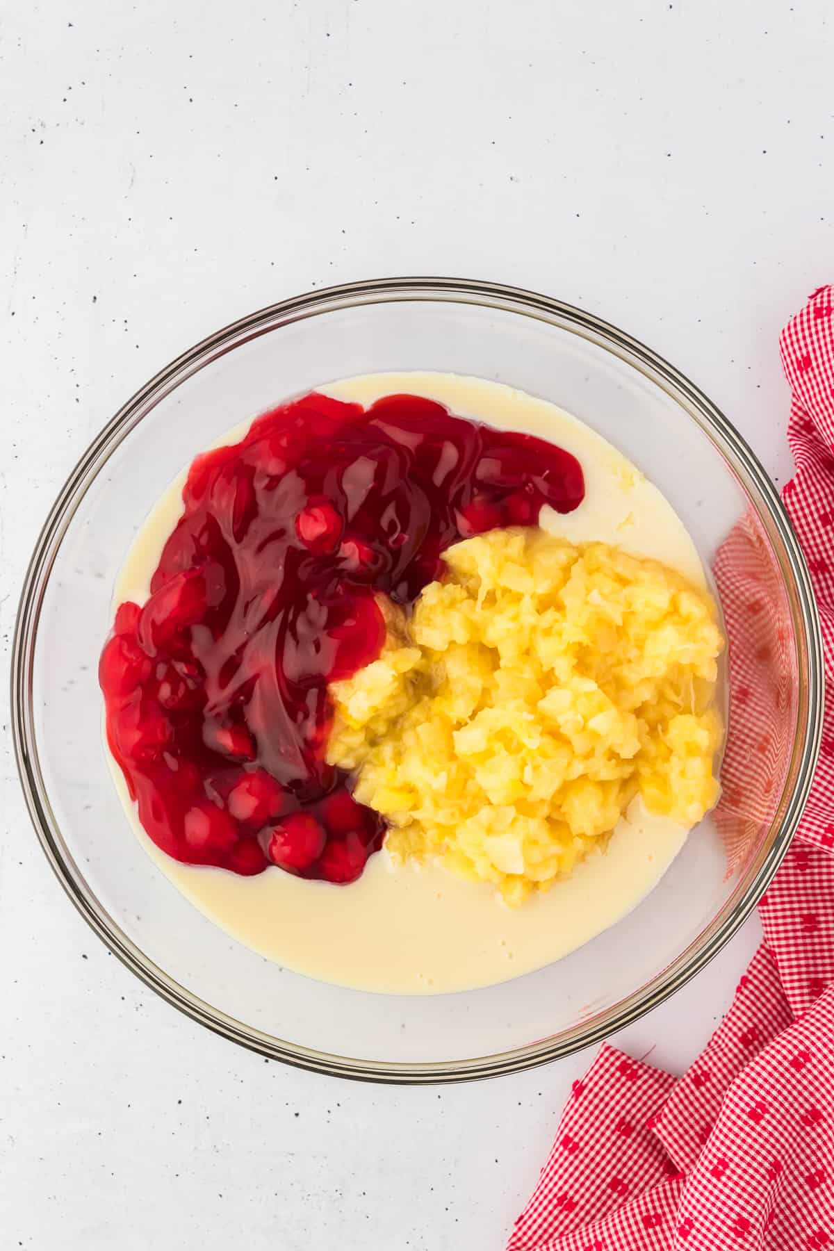 condensed milk, cherry pie filling and crushed pineapple in a clear glass bowl with a red and white kitchen towel to the right of it.