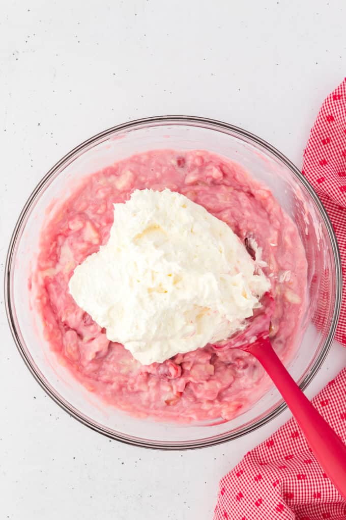 cherry fluff ingredients mixed together with cool whip on top being mixed with a red spatula in a glass bowl with a red and white kitchen towel to the right of it