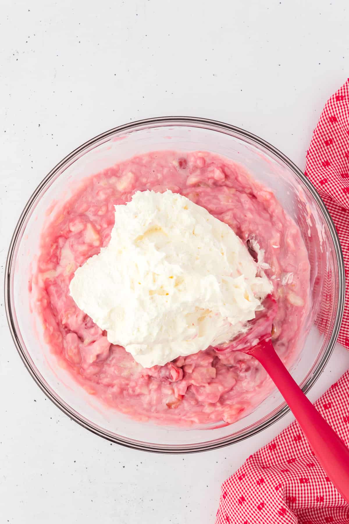 cherry fluff ingredients mixed together with cool whip on top being mixed with a red spatula in a glass bowl with a red and white kitchen towel to the right of it