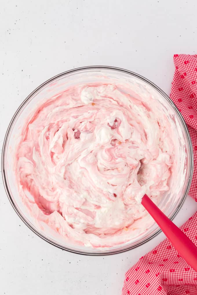 cherry fluff ingredients mixed together with a red spatula in a glass bowl with a red and white kitchen towel to the right of it