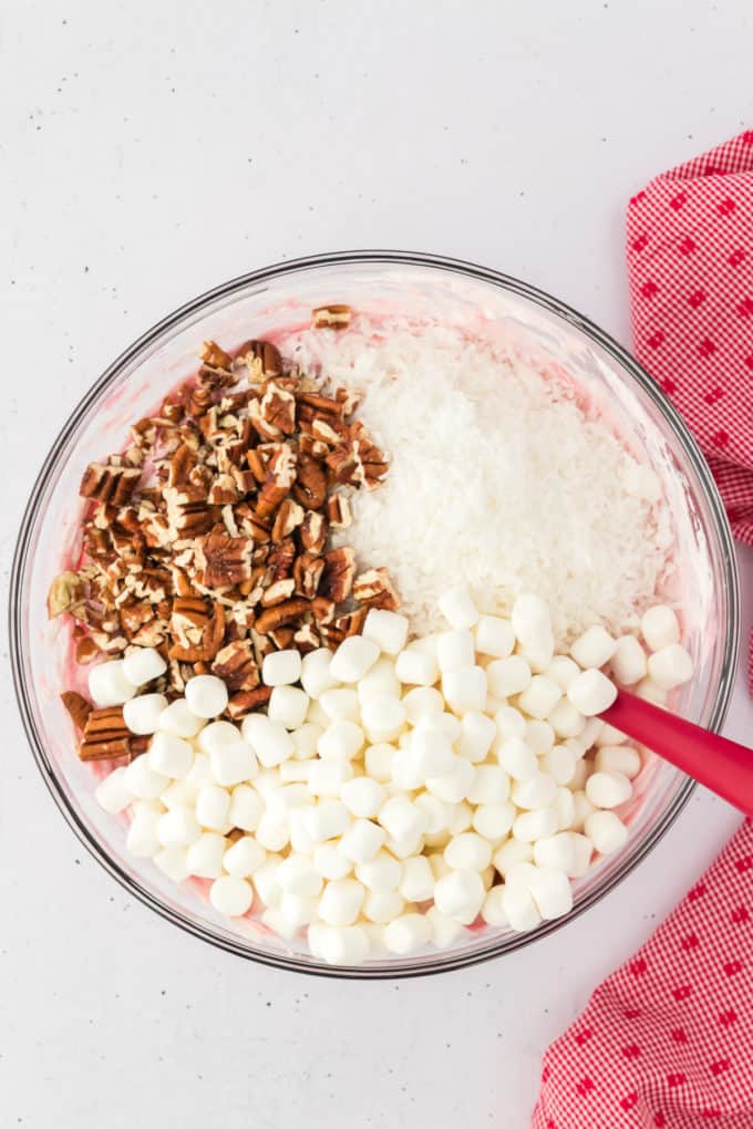 cherry fluff ingredients in a glass bowl with chopped pecans, coconut, and marshmallows on top about to be mixed in with a red spatula, with a red and white kitchen towel to the right of it.