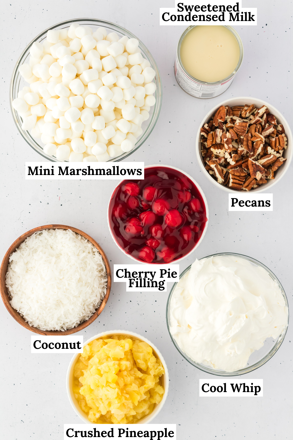 Ingredients for cherry fluff arranged on a flat surface including cherry pie filling, crushed pineapple, sweetened condensed milk, cool whip, sweetened shredded coconut, chopped pecans and mini marshmallows
