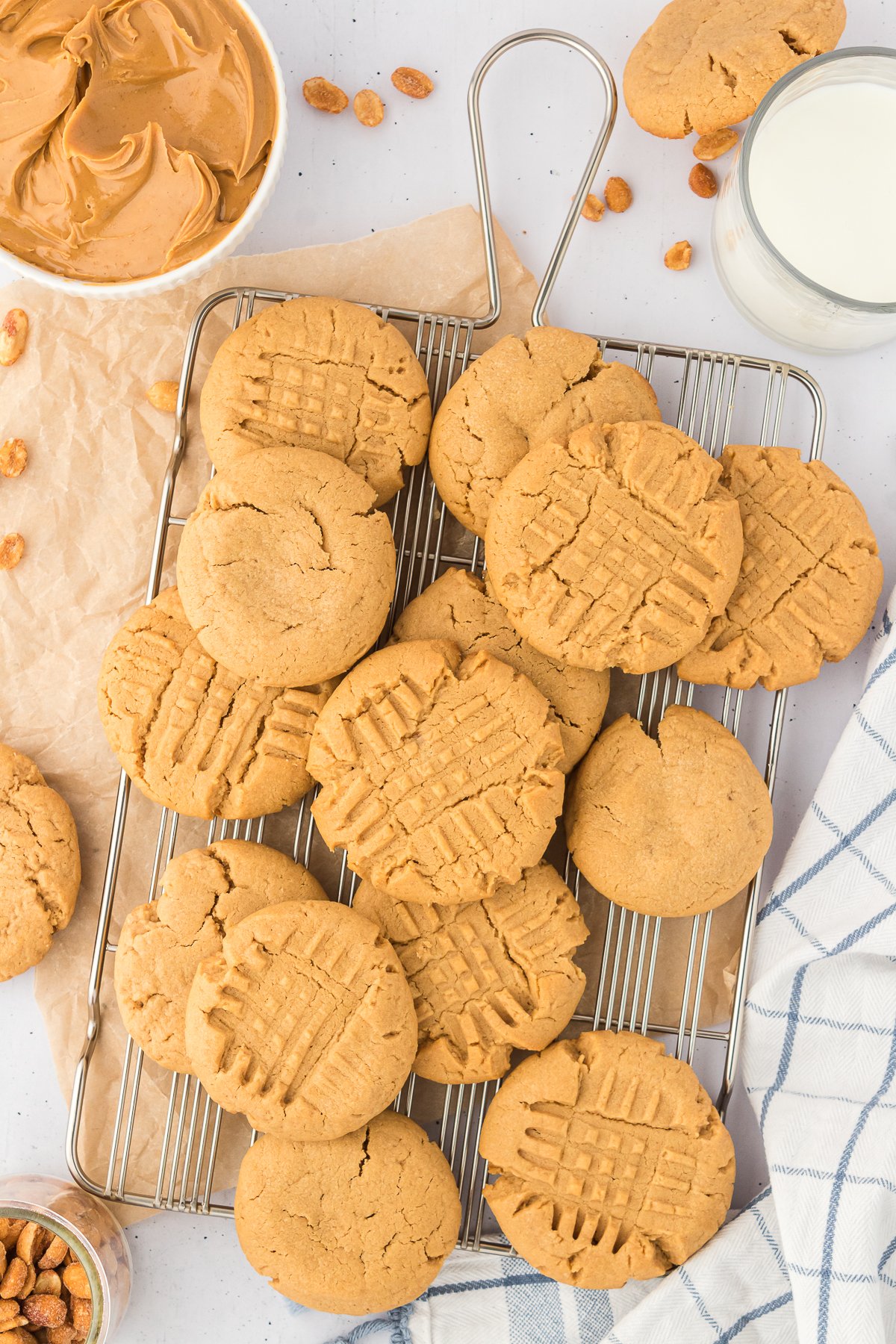 gluten free peanut butter cookies on a wire cooling rack surrounded by a white and blue striped kitchen towel, a bowl of peanuts, a bowl of peanut butter and a glass of milk