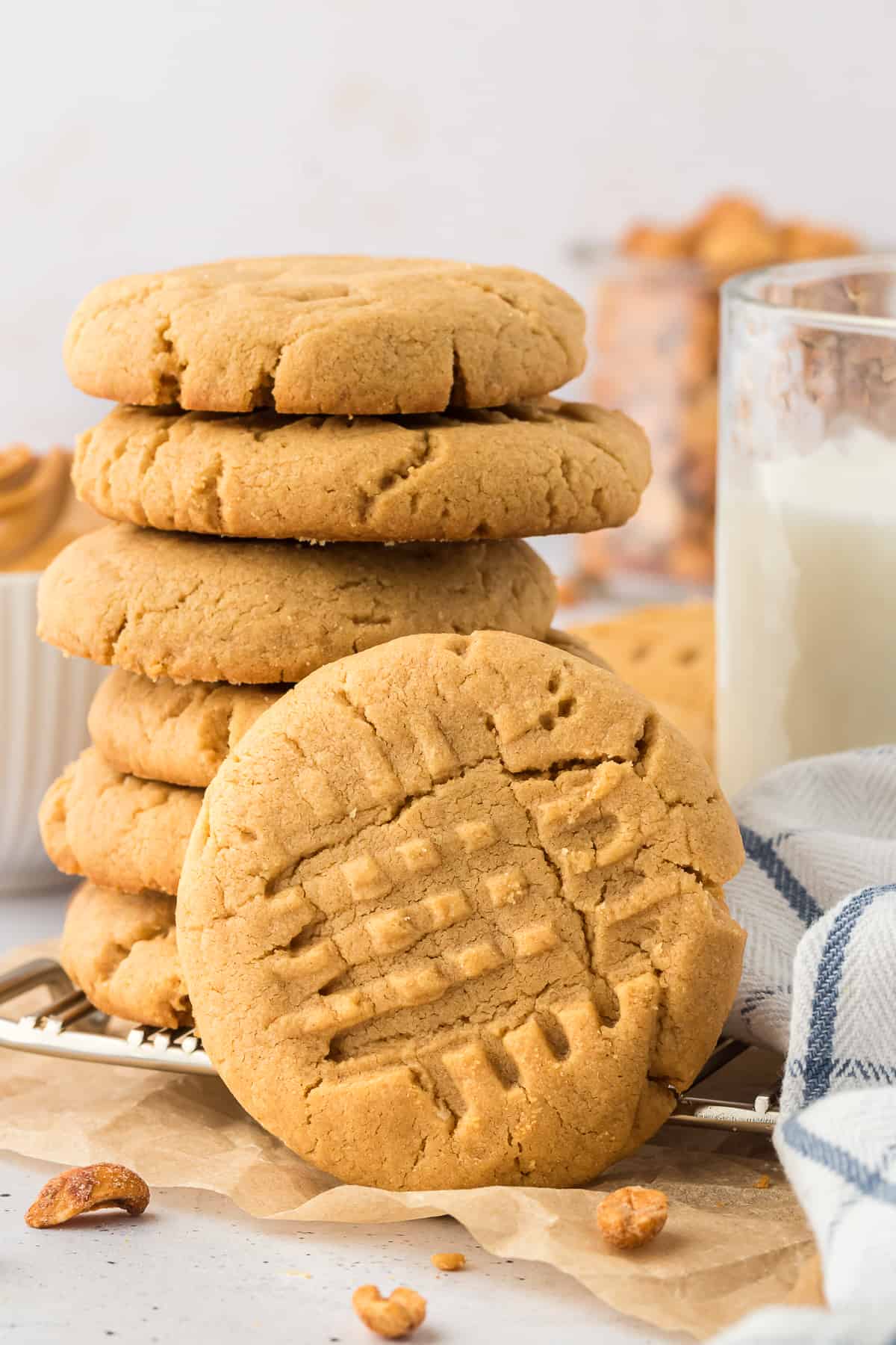 a stack of gluten free peanut butter cookies on a wire cooling rack with one cooking leaning vertically on the side of the stack, a white and blue kitchen towel, peanuts, and a glass of milk around them