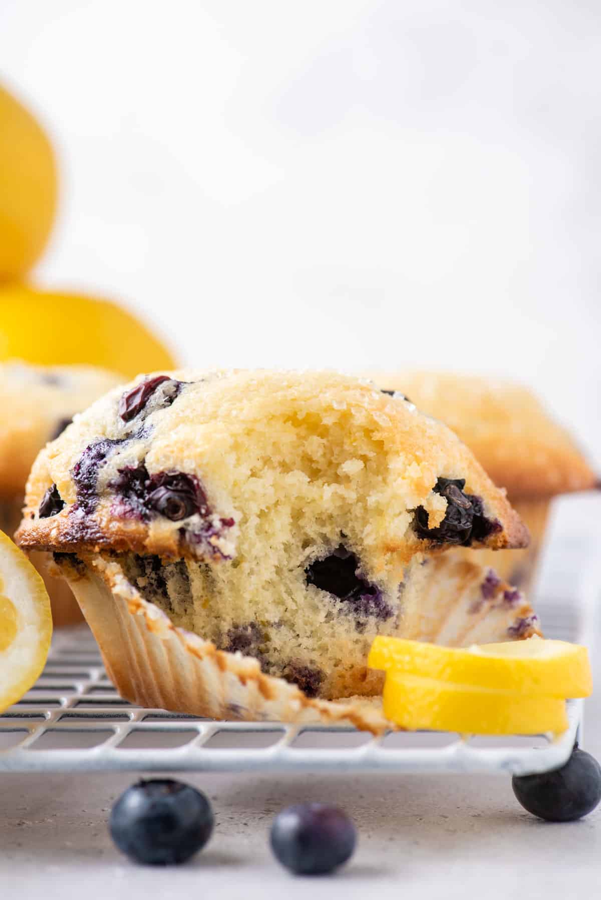 close up of the side of a lemon blueberry muffin with one bite taken out, sitting on top of a wire cooling rack with a lemon peel beside it