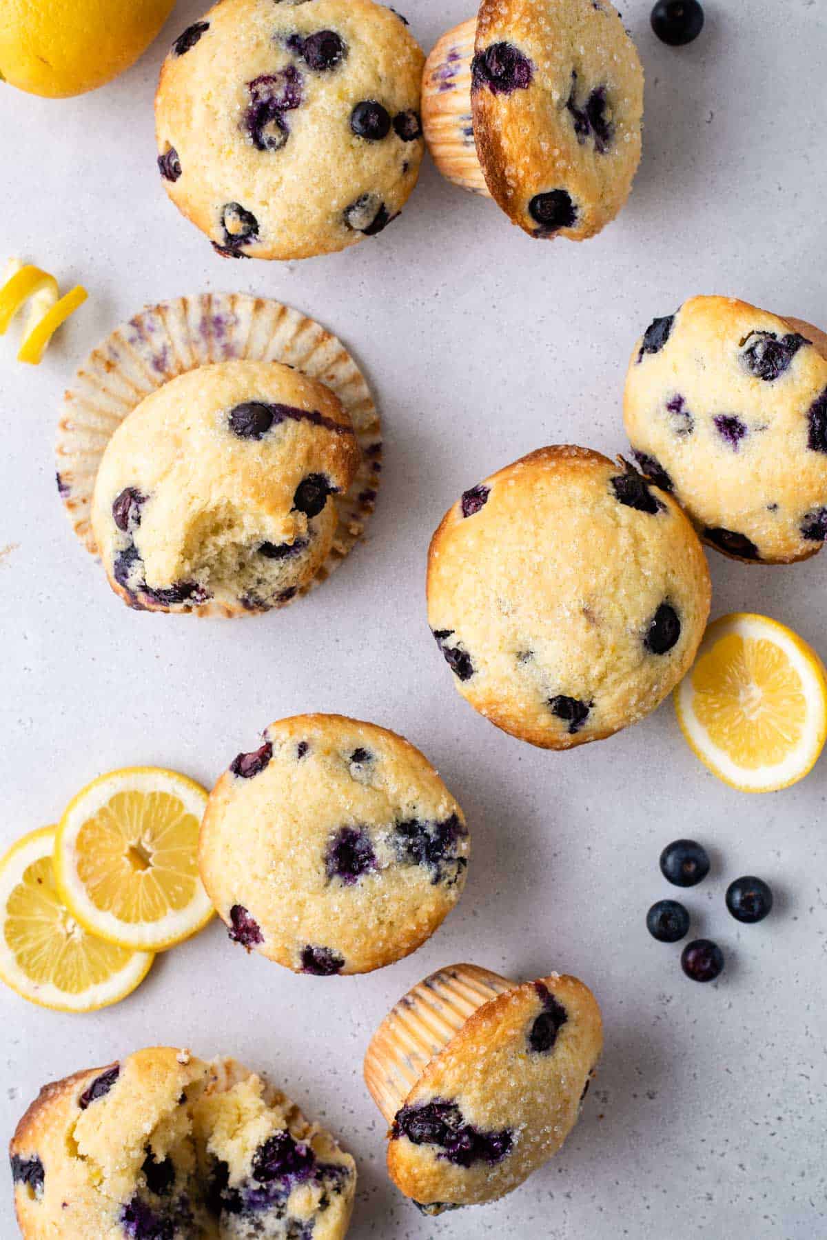 several lemon blueberry muffins arranged on a counter, some whole and some with muffin liners open and bites out, surrounded by lemon slices and whole fresh blueberries