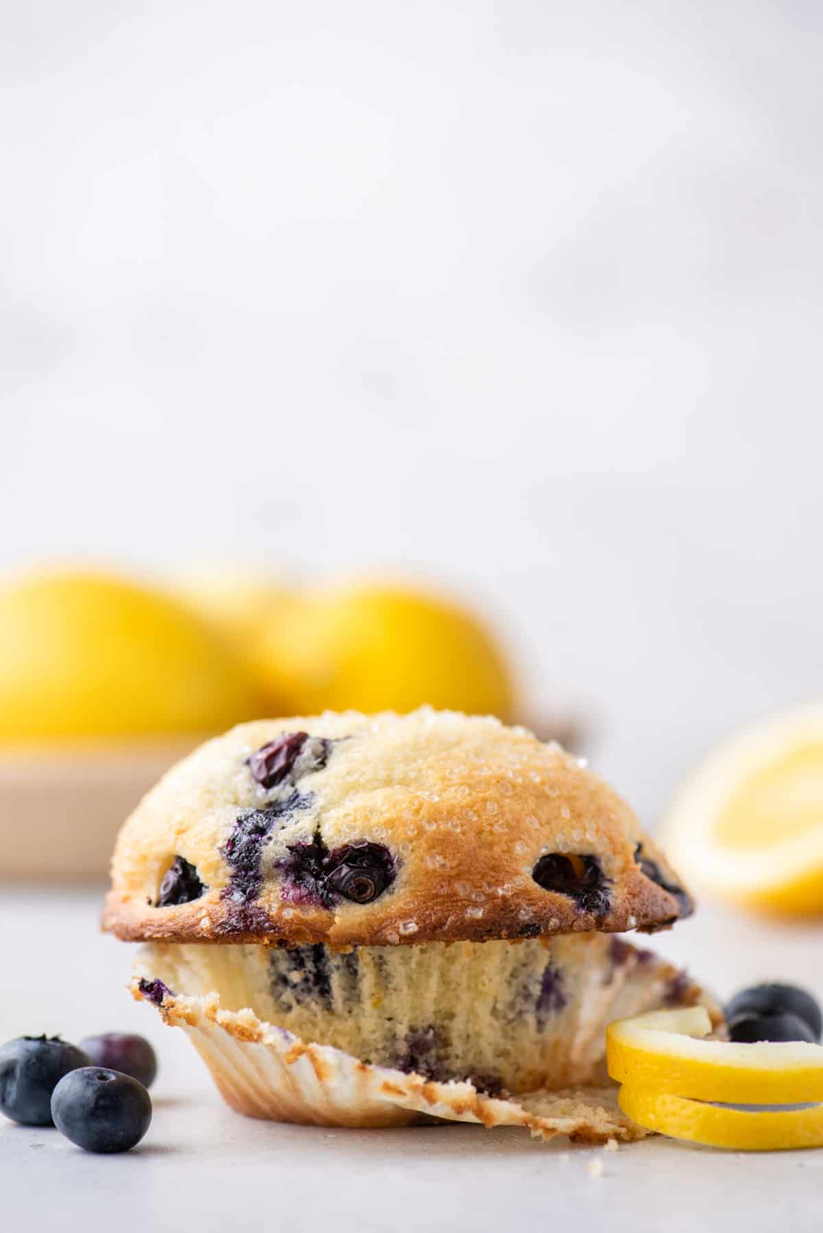 a bakery style lemon blueberry muffin with the muffin liner partially peeled away surrounded by lemon peels and fresh blueberries