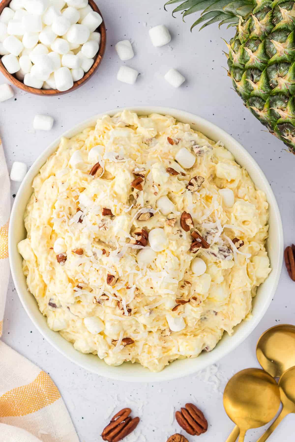 pineapple fluff in a white bowl surrounded by a whole pineapple, a bowl of mini marshmallows, a white and orange kitchen towels, more nuts and scattered mini marshmallows and gold spoons