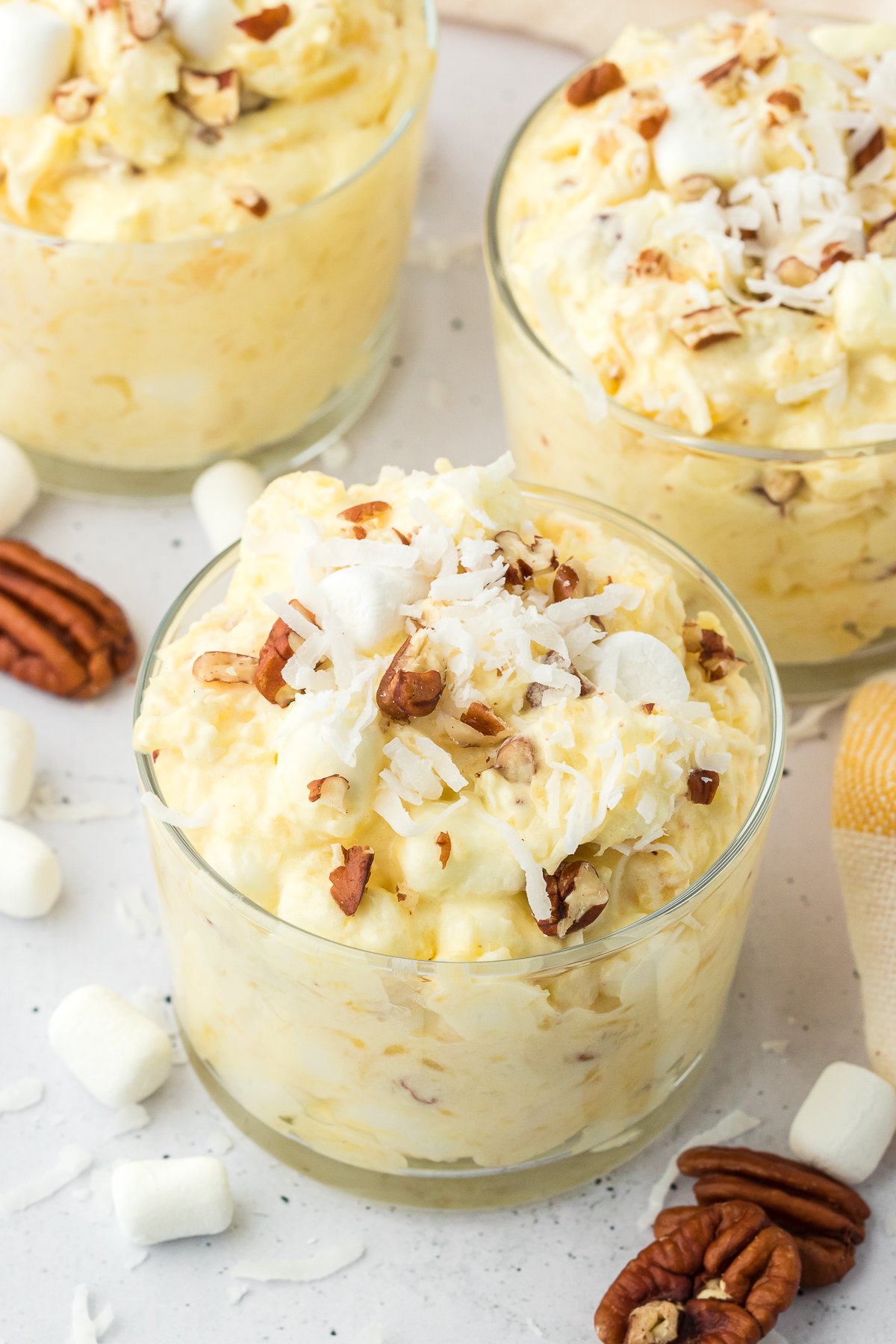 three clear trifle dishes full of pineapple fluff surrounded by nuts, mini marshmallows and shredded coconut
