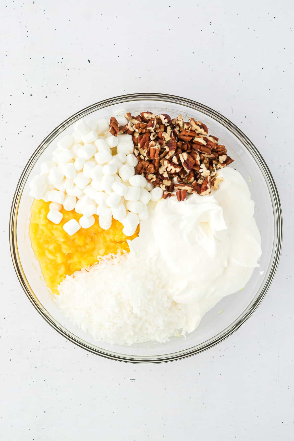 Ingredients for pineapple fluff in a clear glass bowl including crushed pineapple, mini marshmallows, nuts, cool whip and shredded coconut