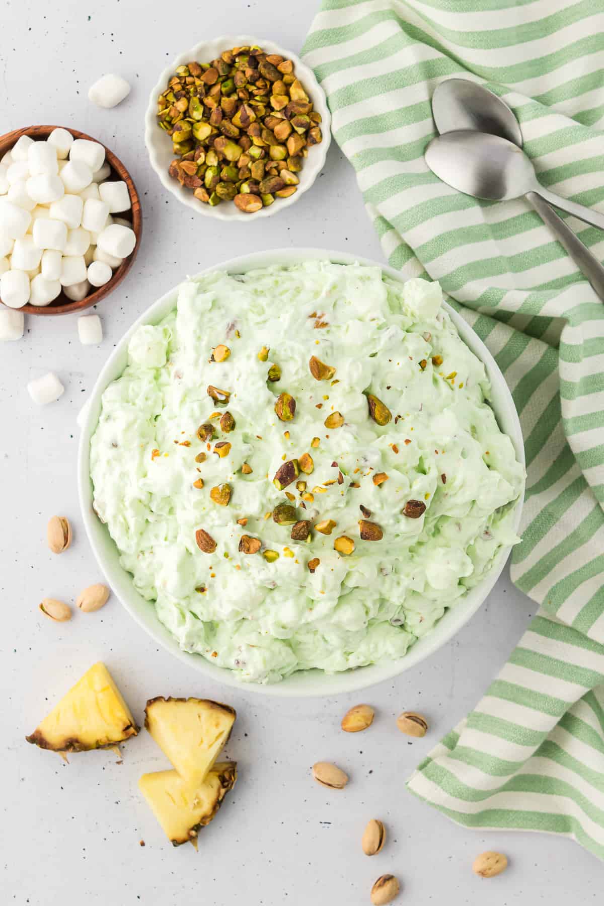 a white bowl full of pistachio salad surrounded by a green and white kitchen towel with two spoons laying on it, a small wooden bowl of mini marshmallows, a small white bowl of pistachios, a few cut pineapple wedges and whole pistachios