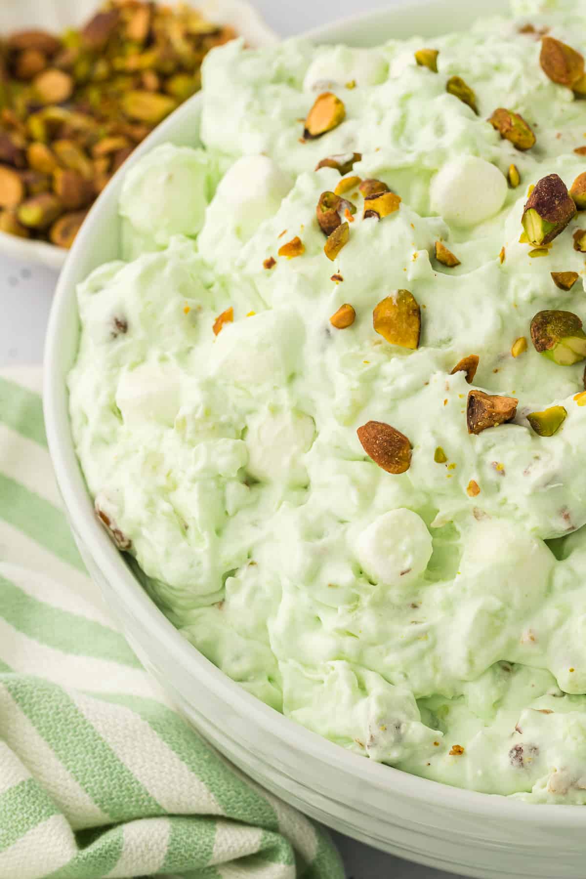 a close up on a large white bowl of pistachio salad with a white and green striped kitchen towel beside it and a bowl of pistachios in the background