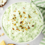 A white bowl filled with light green pistachio salad topped with chopped pistachios, surrounded by mini marshmallows, a striped green towel, and other classic pistachio salad ingredients on a light surface.